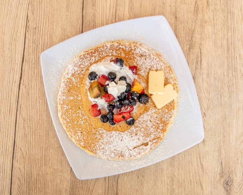 Overhead view of a pancake with whipped cream, blueberries, strawberries, kiwi, and butter.
