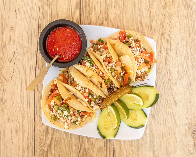 A plate of tacos with salsa, cucumber slices, lime wedges, and a pickled pepper on a wooden table.