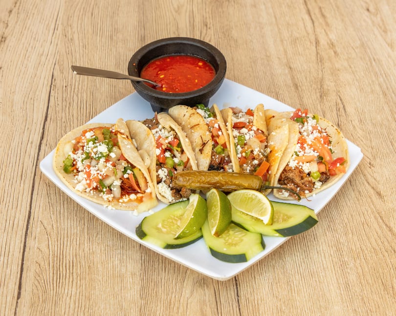 A plate of six tacos with meat, cheese, and salsa, served with limes, cucumbers, and a side of red salsa.