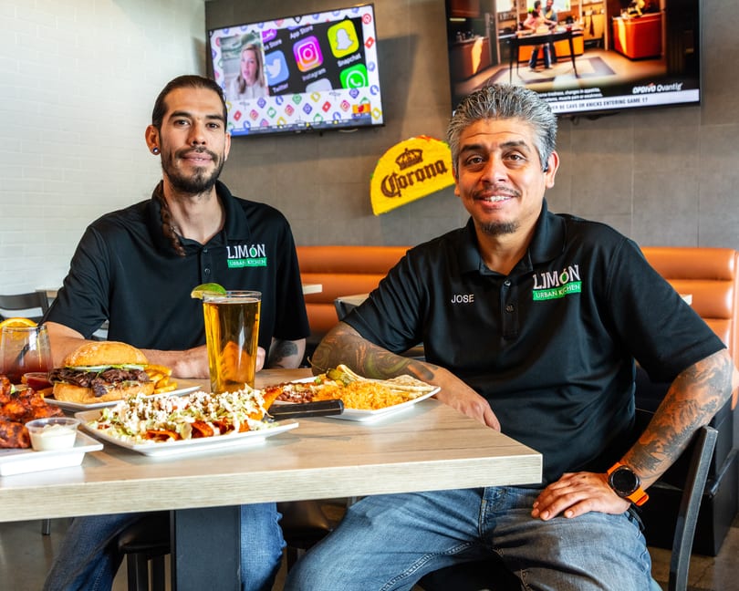 Two men in Limón Urban Kitchen shirts smile at a table laden with food and drinks.