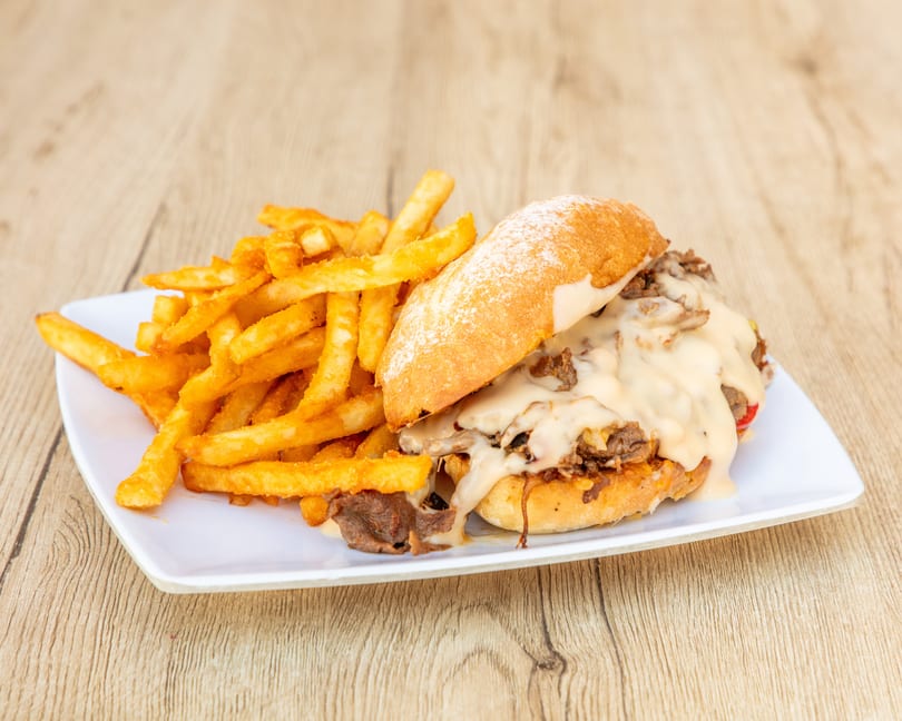 Cheesesteak sandwich with melted cheese and fries on a white plate, on a wooden table.