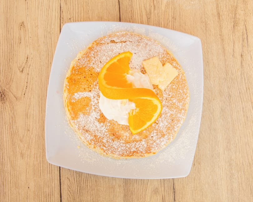 Overhead view of a pancake with powdered sugar, whipped cream, orange slices, and butter on a white plate.